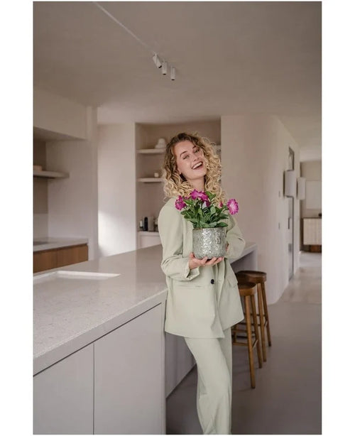 En smilende kvinde med krøllet hår, iført et lyst jakkesæt, står i et moderne, minimalistisk køkken med en Artificial Flower Petunia UV 25 cm mørk pink med levende lyserøde blomster.