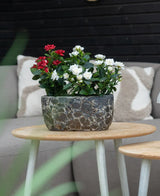 En Artificial Plant Patio Rose 35 cm creme UV står på et rundt træbord foran en beige sofa med mønstrede hynder.