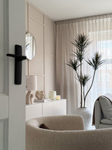 En moderne, minimalistisk stue med en beige tekstureret lænestol, Artificial Plant Dracaena Fragnans 180 cm, hvid skænk med indretning, rundt spejl og transparente gardiner, der filtrerer blødt dagslys gennem vinduet.