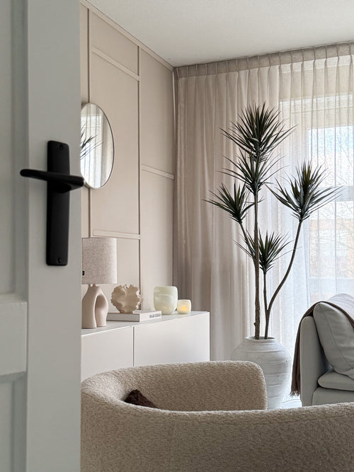 En moderne, minimalistisk stue med en beige tekstureret lænestol, Artificial Plant Dracaena Fragnans 180 cm, hvid skænk med indretning, rundt spejl og transparente gardiner, der filtrerer blødt dagslys gennem vinduet.