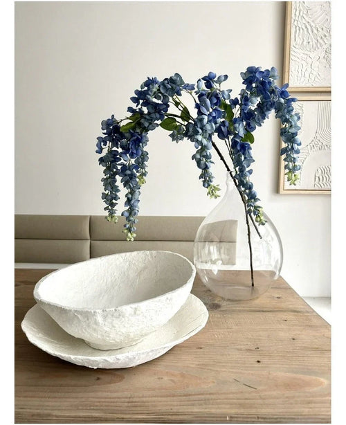 Et blomsterarrangement fra Kunst Wisteria på 115 cm står i en gennemsigtig vase på et træbord ved siden af to hvide skåle med struktur, mens væggen bagved er dekoreret med abstrakt kunst.