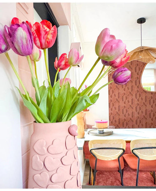 En vase med lyserøde hjertemotiver holder Kunstig Blomst Fransk Tulipan 60 cm lilla på et spisebord med stole med vævet ryglæn. I baggrunden bidrager et rundt spejl og en brunmønstret væg til det hyggelige, moderne interiør.