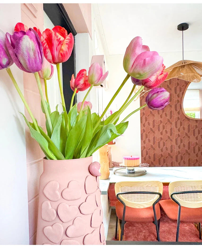 Een roze vaas met hartjes in reliëf houdt de Kunstbloem Franse Tulp 60 cm rood op een eettafel. Daarachter staan stoelen met rieten ruggen, een taart op tafel en een spiegel op een gedessineerde bruine muur onder een geweven hanglamp.