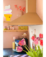 Et pastellyserødt køkken med Artificial Flower French Tulip 60 cm lilla i forgrunden, en gul LOVETTI tallerken på hylden, lyserøde krus, vægkunst af en iskugle og et minimalistisk vægmaleri af to personer på væggen.