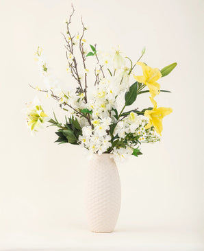 En vase med en hvid struktur viser elegancen af Sunkissed Ivory kunstige blomsterbuket med gule liljer, hvide blomster og grønne blade mod en almindelig lys baggrund.