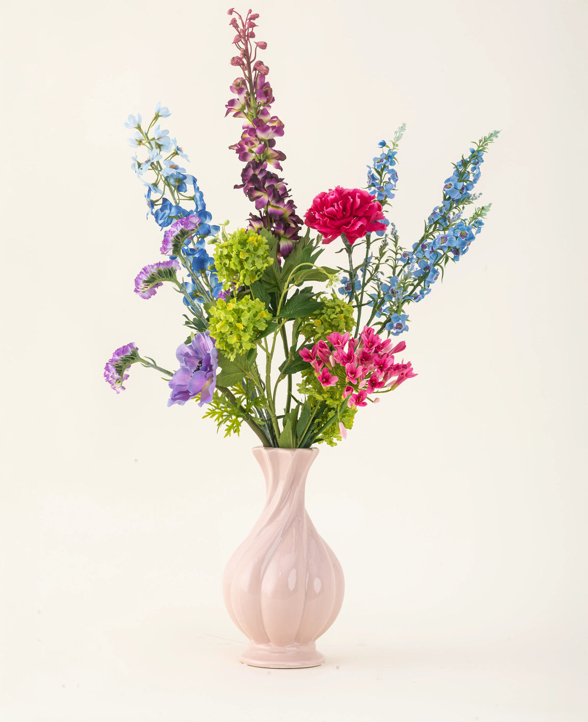 Arrangeret i en pink keramisk vase, Blossom Breeze kunstige blomsterbuket i silke byder på livlige blå, lilla, røde og grønne blomster - en vedligeholdelsesfri blomsterbuket mod en almindelig lys baggrund.
