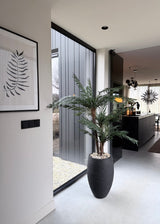 Et moderne, minimalistisk interiør med en Artificial Cycas Classic Palm 135 cm i en stilfuld dekorativ potte nær et gulv til loft-vindue, et bregnekunstværk på væggen og et lækkert sort køkken med hængende lamper i baggrunden.