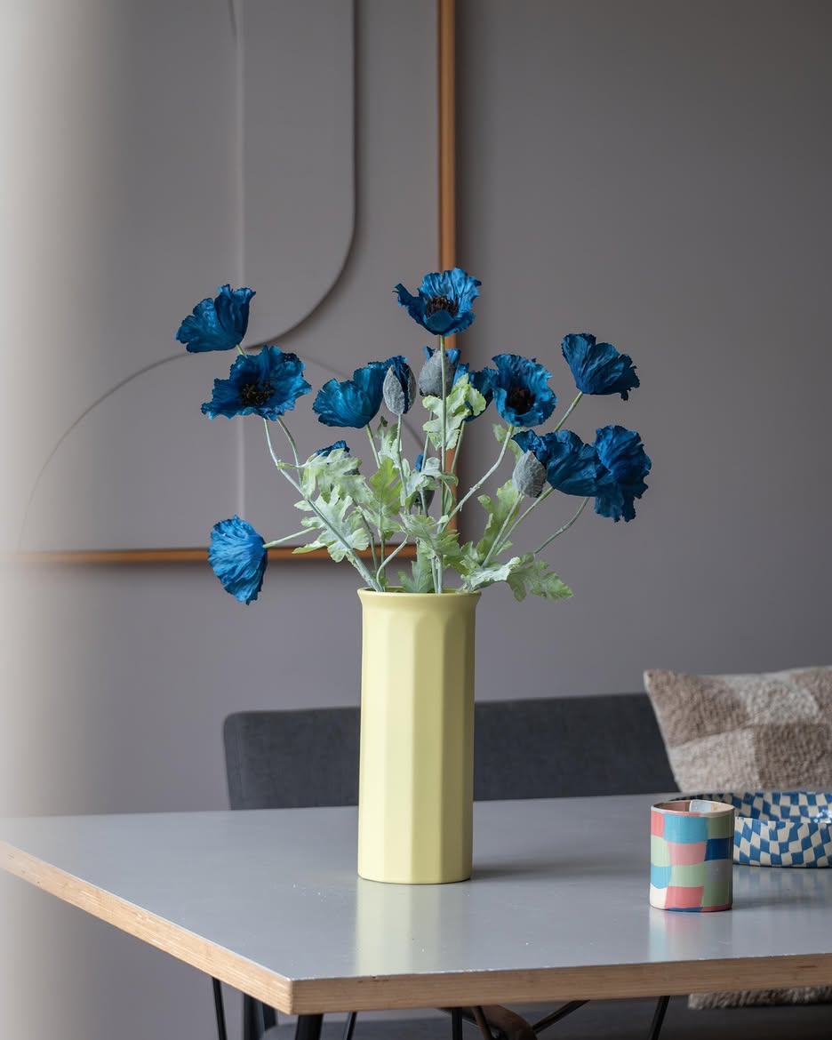 En 73 cm kunstig blomstervalmue i mørkeblå står i en gul vase på et bord, ledsaget af en lille, farverig stribet kop, i et moderne, blødt oplyst rum med minimalistisk indretning og neutrale toner.