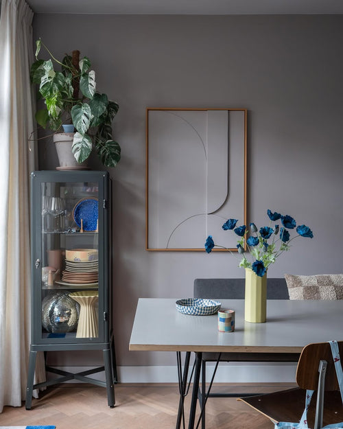Den moderne spiseplads har et gråt vitrineskab, hvor service og planter er udstillet, minimalistisk vægdekoration og et bord, hvorpå Kunstig Blomstervalmue 73 cm mørkeblå står i en gul vase. Rummet fuldendes af en eklektisk blanding af stole, alt sammen mod en baggrund af grå vægge og et trægulv.