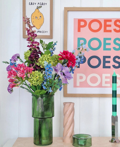 En grøn glasvase holder en kunstig silkeblomstbuket Blossom Breeze på bordet. Bag den hænger indrammede billeder - et med en citron og "EASY PEASY", et andet med "POESY" - hvilket tilføjer ekstra charme. I nærheden er et grønt lys og et pink spirallys fra Blossom Breeze.