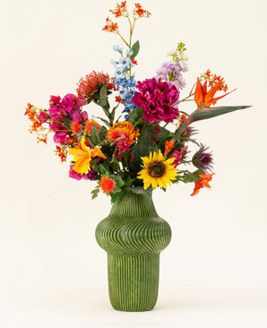 En grøn struktureret vase viser Colorful Spirit kunstige blomsterbuket i silke med solsikker, lyserøde pæoner, orange og lilla blomster og frodigt grønt mod en almindelig lys baggrund.