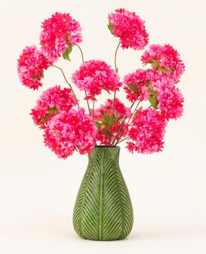 En grøn vase med bladmotiv viser den kunstige silkeblomstbuket Cherry Blossom Pink - lys pink, luftige silkekirsebærblomster med grønne blade - mod en almindelig lys baggrund.