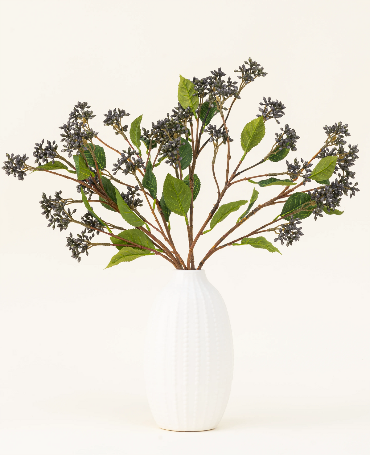 En kunstig silkeblomstbuket Japanese Berry Steel Blue med lange stilke, mørkelilla bær og stålblade står i en vase med hvid tekstur mod en almindelig lys baggrund.