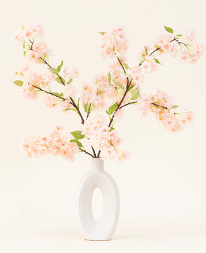 En hvid keramikvase med en bred oval åbning viser flere grønbladede kunstige silkeblomsterbuketter Kirsebærblomster lyserøde grene mod en enkel lys baggrund.