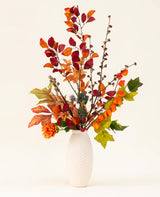 En hvid tekstureret vase fremviser Chestnut Charm Artificial Flower Bouquet, med efterårsblade, orange blomster og grønt løv - et elegant kunstigt arrangement mod en almindelig lys baggrund.