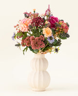 I en hvid, buet keramikvase er Pretty Pastel kunstige blomsterbuket i silke - en farverig blanding af bæredygtige blomster såsom lyserøde roser, georginer, lilla og orange blomster og grønt - smukt arrangeret mod en lys baggrund.