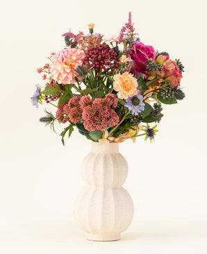 I en hvid, buet keramikvase er Pretty Pastel kunstige blomsterbuket i silke - en farverig blanding af bæredygtige blomster såsom lyserøde roser, georginer, lilla og orange blomster og grønt - smukt arrangeret mod en lys baggrund.