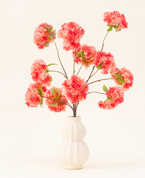 Silk Artificial Flowers Bouquet Cherry Blossom Coral har levende lyserøde og kunstige koralkirsebærblomster med grønne blade, elegant arrangeret i en hvid skulptureret vase mod en lys baggrund.
