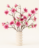 Silke kunstige blomsterbuketten Magnolia pink er arrangeret i en bølget hvid keramikvase med grønne knopper, hvilket skaber et elegant display mod en lys baggrund.