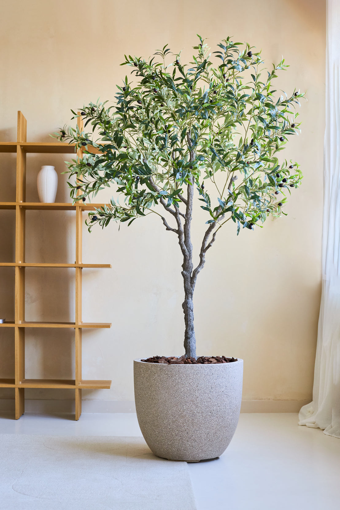 Een Kunst Olijfboom 200 cm kunstplant met slanke takken en groene bladeren staat op een lichte vloer bij een houten plank en transparante gordijnen in een lichte, minimalistische kamer.