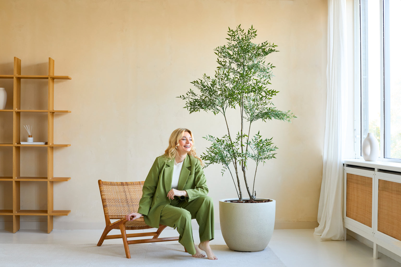 En kvinde i grønt jakkesæt smiler, mens hun sidder på en træstol ved siden af Artificial Ruscus 180 cm planten i et lyst, moderne rum med beige vægge, store vinduer og minimalistisk indretning.