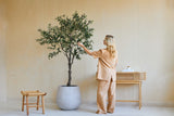 En kvinde i løst beige tøj står ved siden af et 200 cm kunstigt oliventræ og rører blidt ved bladene. I nærheden står en træskammel og et konsolbord med bøger og en diffuser op ad den beige væg.