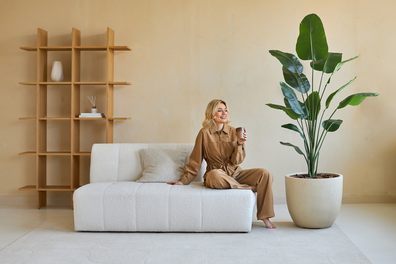 En kvinde i brunt outfit sidder på en hvid sofa og smiler med en kop i hånden. Ved siden af hende er den kunstige plante Strelitzia 180 cm ægte touch, som bringer frodigt grønt ind i den moderne, beige-tonede stue med minimal indretning.