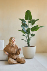 En kvinde i brun jumpsuit sidder smilende på gulvet med en kaffekop ved siden af, ved siden af den kunstige plante Strelitzia 180 cm ægte touch i et minimalistisk rum med beige vægge.