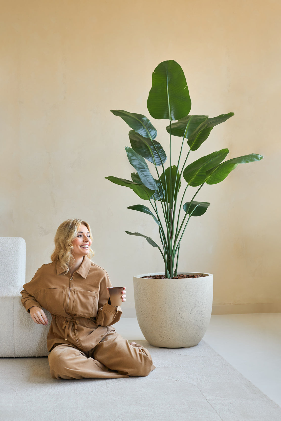En kvinde i brun jumpsuit sidder smilende på gulvet med en kaffekop ved siden af, ved siden af den kunstige plante Strelitzia 180 cm ægte touch i et minimalistisk rum med beige vægge.