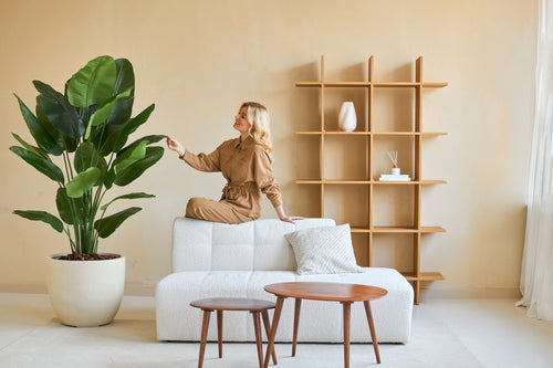 En kvinde i brun jumpsuit sidder på ryggen af en hvid sofa og rører ved bladet på en Artificial Strelitzia Real Touch Deluxe 180 cm plante i en minimalistisk stue med træborde og hylder.