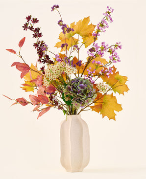 'Wild Ametyst'-buketten af kunstige silkeblomster, med efterårsblade, lilla og hvide blomster og grønne hortensiaer, er i en hvid keramikvase mod en almindelig, lys baggrund.