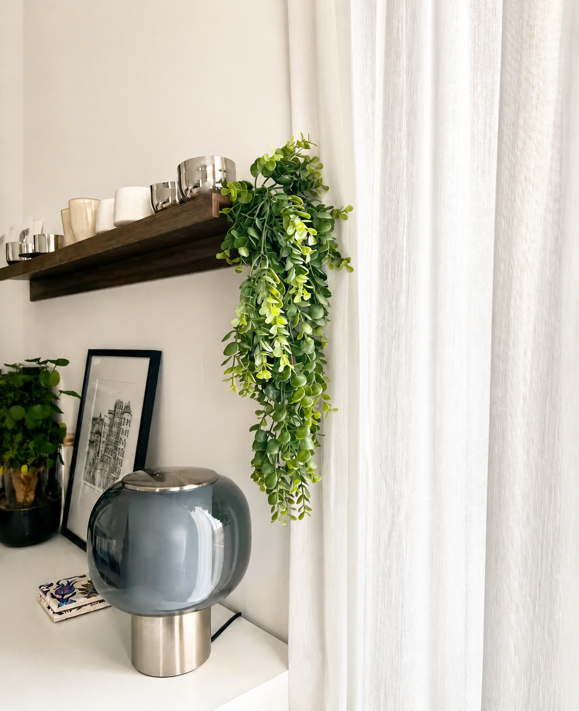 Et moderne opholdsrum med en grå glaslampe, indrammet kunst, en potteplante og en træhylde med kopper og den kunstige hængeplante 'Pannenkoek' (61 cm), hvis ranker hænger ved siden af hvide gardiner.