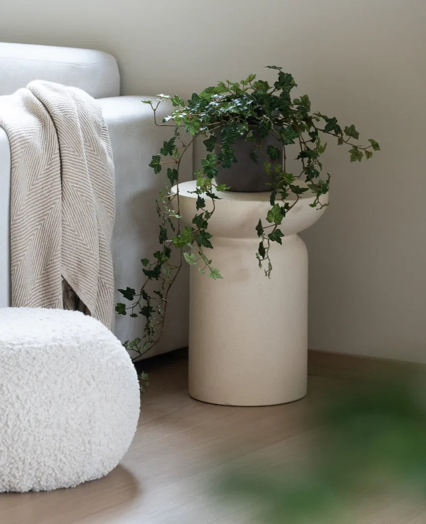 En Artificial Ivy Deluxe Green 90 cm står i en beige vase på et trægulv ved siden af en lysegrå sofa med en cremefarvet plaid og en hvid struktureret puf.