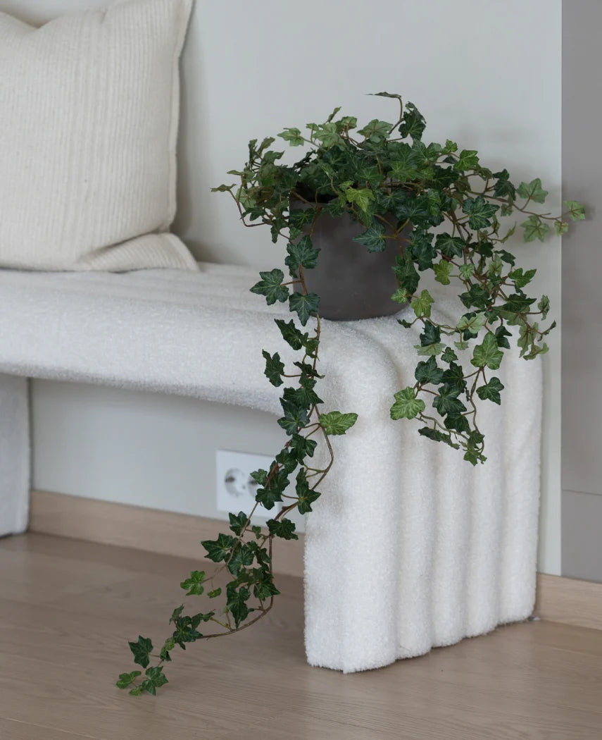 De Kunst Klimop Deluxe Groen 90 cm, met zijn weelderige hangende ranken, staat op een wit bankje met structuur naast een crèmekleurig kussen in een minimalistische kamer met lichte houten vloeren.