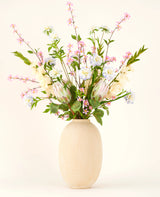 I en vase med en beige struktur ligger Silk kunstig blomsterbuket Lots of Love XL, en håndbundet buket af lyserøde, hvide og blå silkeblomster med grønne blade og lange stilke mod en lys baggrund.