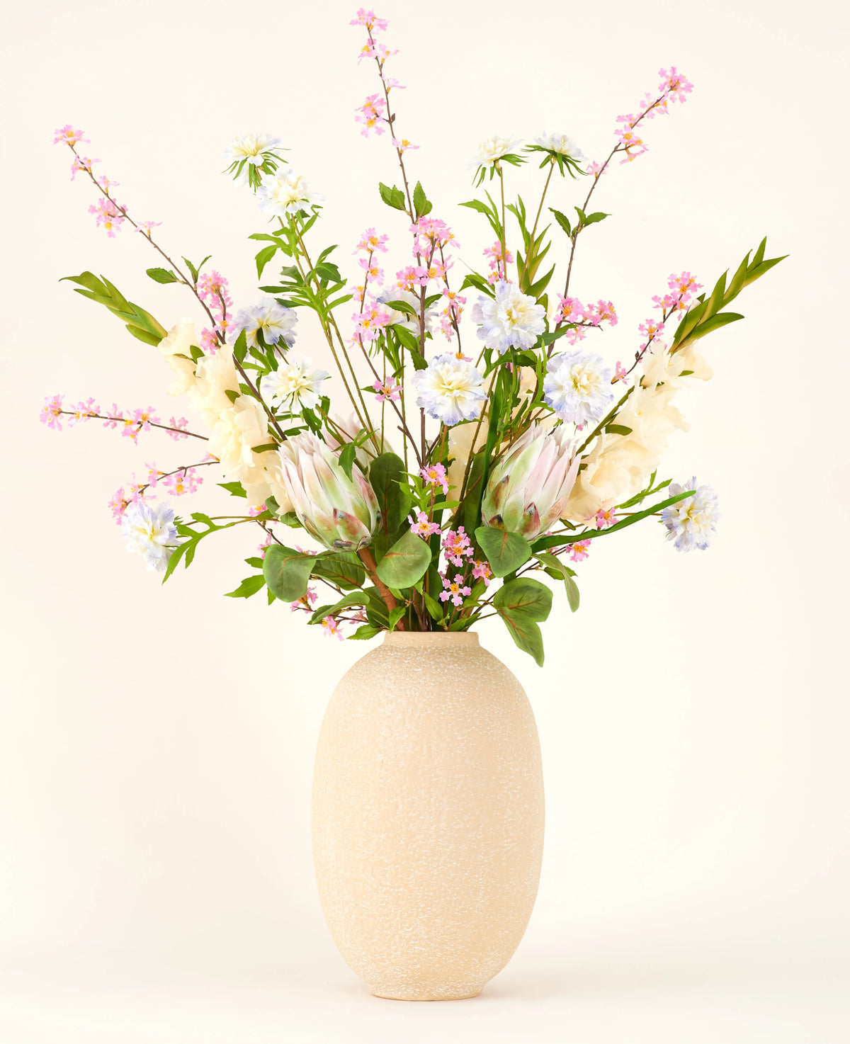 I en vase med en beige struktur ligger Silk kunstig blomsterbuket Lots of Love XL, en håndbundet buket af lyserøde, hvide og blå silkeblomster med grønne blade og lange stilke mod en lys baggrund.