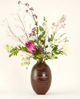 En Serene Bloomfield kunstig blomsterbuket i silke, med en stor pink protea, hvide roser, lilla og hvide blomster og grønt løv, står i en brun ribbet vase mod en almindelig lys baggrund.