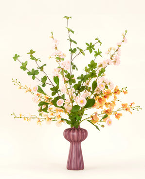En Blossom Harmony kunstig blomsterbuket i silke er arrangeret i en lilla vase med lyserøde og ferskenblomster og grønne blade på en enkel lys baggrund for en elegant visning.