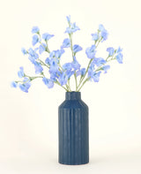En blå ribbet keramikvase viser Pea Blue kunstige blomsterbuket i silke med sine sarte lyseblå kunstige blomster mod en almindelig, lys baggrund.