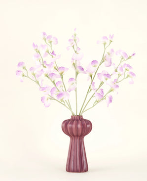 Silke kunstige blomsterbuketten Pea lilac har lange stængler af sarte, lyse lilla blomster og grønne blade i en lilla keramisk vase med lodrette kanter, mod en almindelig, råhvid baggrund.