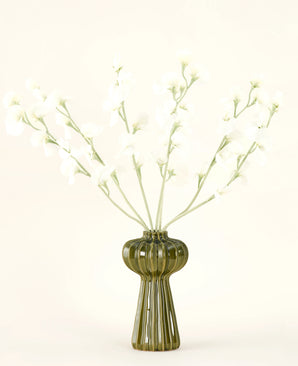 En grøn ribbet glasvase holder Silk kunstige blomsterbuket Ærtecreme med sarte hvide silkeblomster og slanke stilke, mod en almindelig råhvid baggrund.