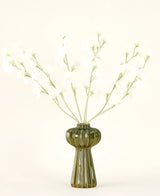 En grøn ribbet glasvase holder Silk kunstige blomsterbuket Ærtecreme med sarte hvide silkeblomster og slanke stilke, mod en almindelig råhvid baggrund.