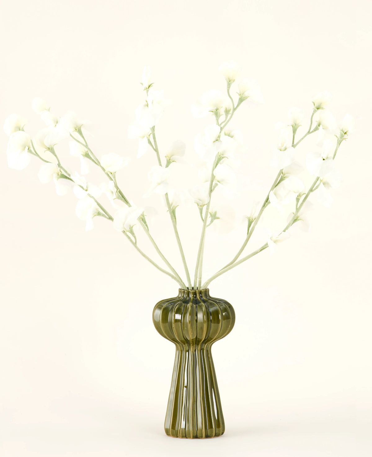 En grøn ribbet glasvase holder Silk kunstige blomsterbuket Ærtecreme med sarte hvide silkeblomster og slanke stilke, mod en almindelig råhvid baggrund.
