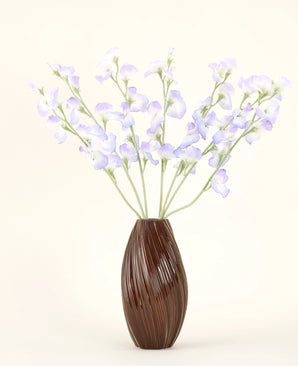 En brun, ribbet vase holder den kunstige silkeblomstbuket Ærtelavendel med sarte kronblade og grønne stængler mod en almindelig råhvid baggrund for en subtil sommerhusatmosfære.