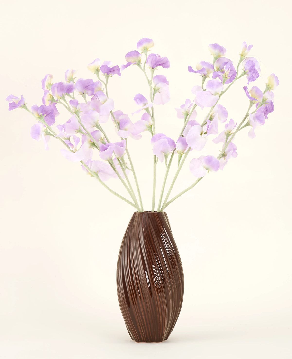 En tekstureret brun vase holder den kunstige silkeblomstbuket Ært lilla-lyse lilla silkeblomster med grønlige stilke mod en almindelig, lys baggrund.