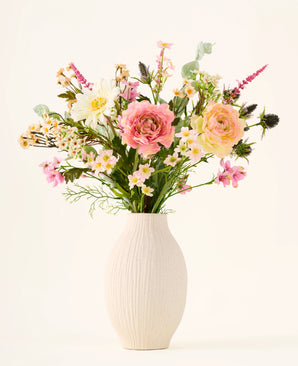 I en hvid keramikvase er 'Backyard Blossom'-buketten af kunstige silkeblomster, bestående af et smukt arrangement af lyserøde roser, hvide tusindfryd og små gule blomster, lavet af silkeblomster, suppleret med frodige grønne blade mod en almindelig lys baggrund.