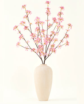 En høj beige keramikvase indeholder flere grene fra Silke kunstige blomsterbuketten Pink blomster, med sarte lyserøde blomster og grønne blade, mod en almindelig hvid baggrund.