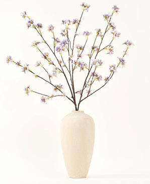 En høj, cremefarvet keramikvase indeholder en række tynde grene med teksten Silke kunstig blomsterbuket Kunstig blomstergren lilla, mod en almindelig, lys baggrund.