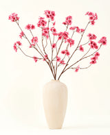 Silke kunstige blomsterbuketten Cherry blossom fuchsia 114 cm tilføjer en levende pop af farve til dit interiør med sine slående grene og fuchsia blomster, smukt udstillet i en beige keramisk vase mod en almindelig råhvid baggrund.