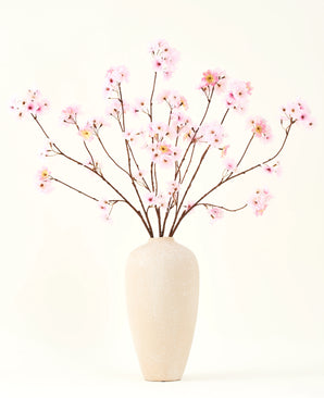 Silke kunstige blomsterbuketten Cherry blossom pink 114 cm har lange, tynde grene med bundter af lyserøde blomster i en beige keramikvase, som danner en slående kontrast mod den almindelige hvide baggrund.